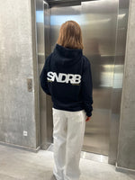Stemningsbillede af pige der bærer en navy hoodie med hvidt SNDRB tryk og hvide NANNA jeans.  Fra BySofieSønderby