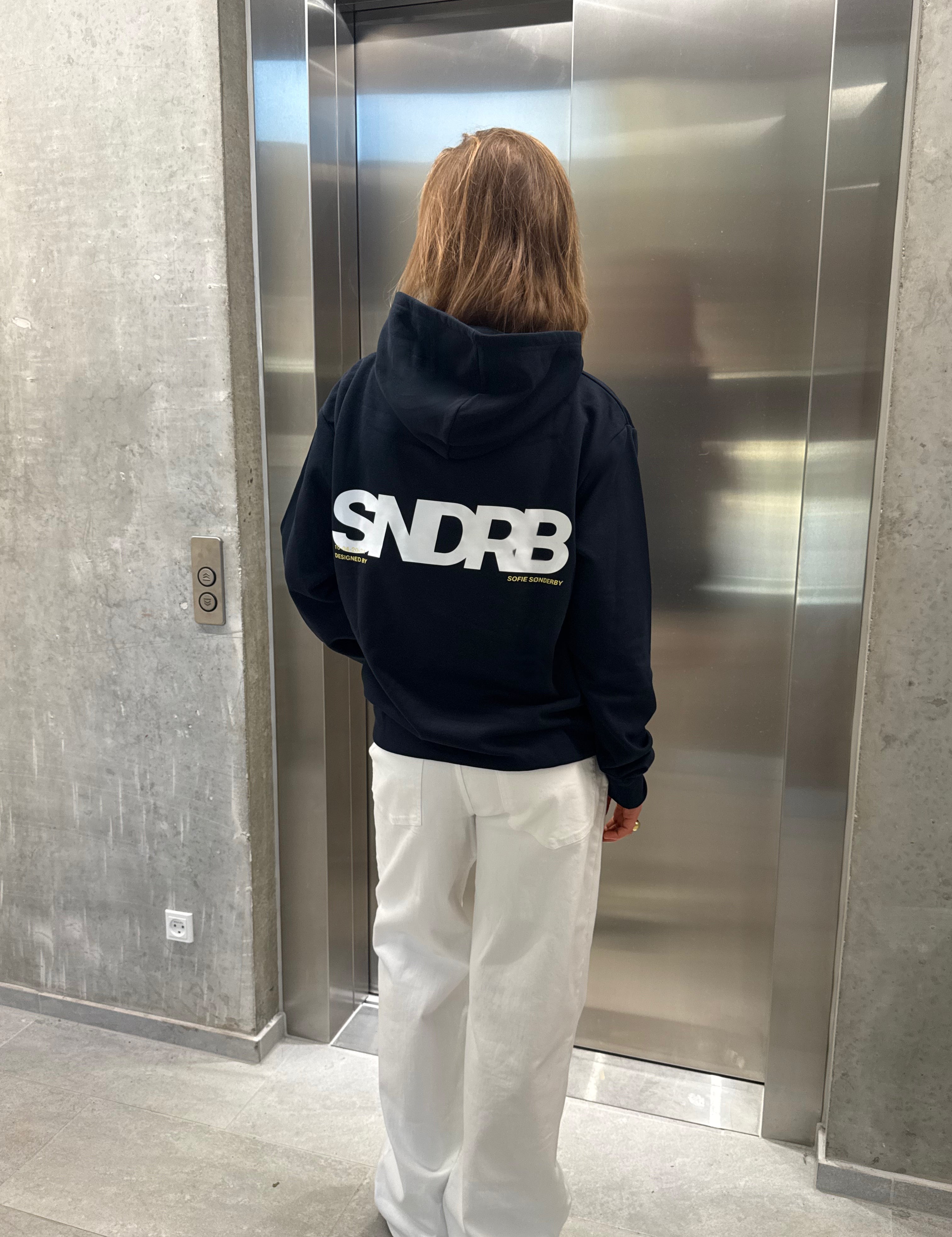Stemningsbillede af pige der bærer en navy hoodie med hvidt SNDRB tryk og hvide NANNA jeans.  Fra BySofieSønderby