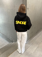 Stemningsbillede af pige der bærer en sort hoodie med gult SNDRB tryk og hvide NANNA jeans.  Fra BySofieSønderby