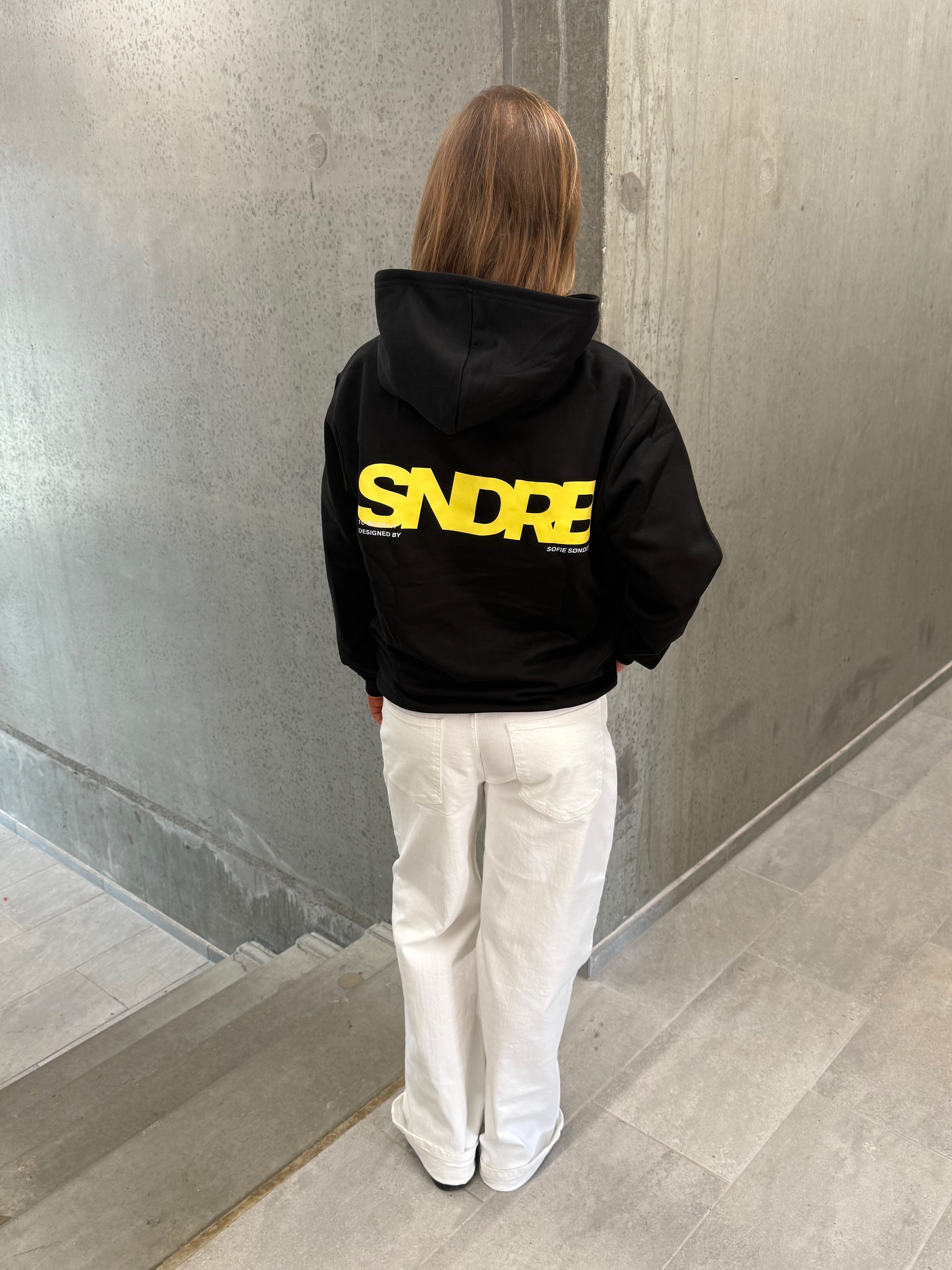 Stemningsbillede af pige der bærer en sort hoodie med gult SNDRB tryk og hvide NANNA jeans.  Fra BySofieSønderby