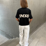 Stemningsbillede af pige der bærer en sort t-shirt med beige SNDRB tryk og hvide NANNA jeans.  Fra BySofieSønderby