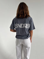 Modelfoto af pige som bærer en SNDRB tee i farven stone, samt et par hvide jeans. Alt fra By Sofie Sønderby