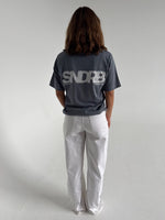 Modelfoto af pige som bærer en SNDRB tee i farven stone, samt et par hvide jeans. Alt fra By Sofie Sønderby