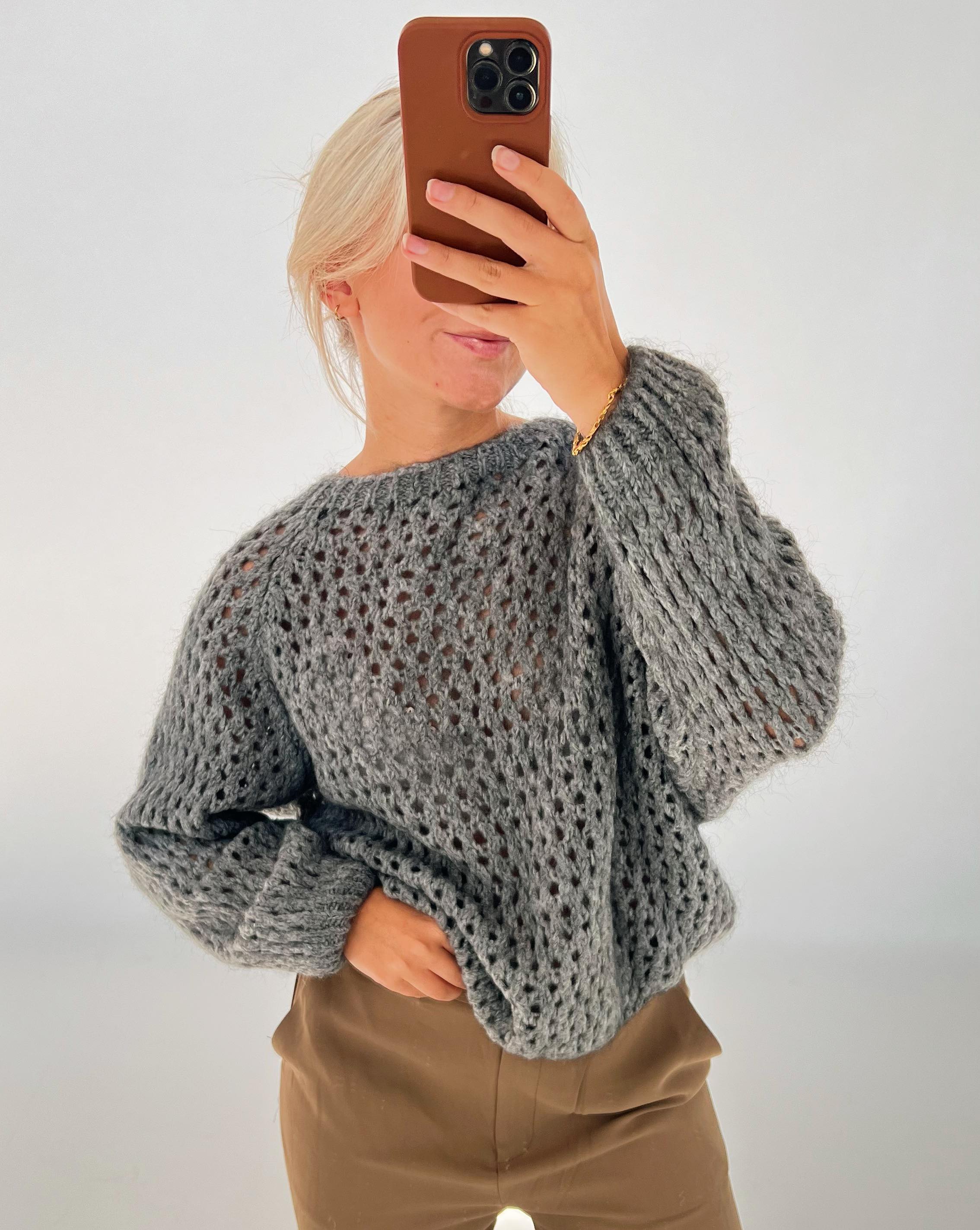 En person tager en spejlslefie med en mobiltelefon, iført en grå, strikket sweater og brune bukser. Baggrunden er simpel og lys.
