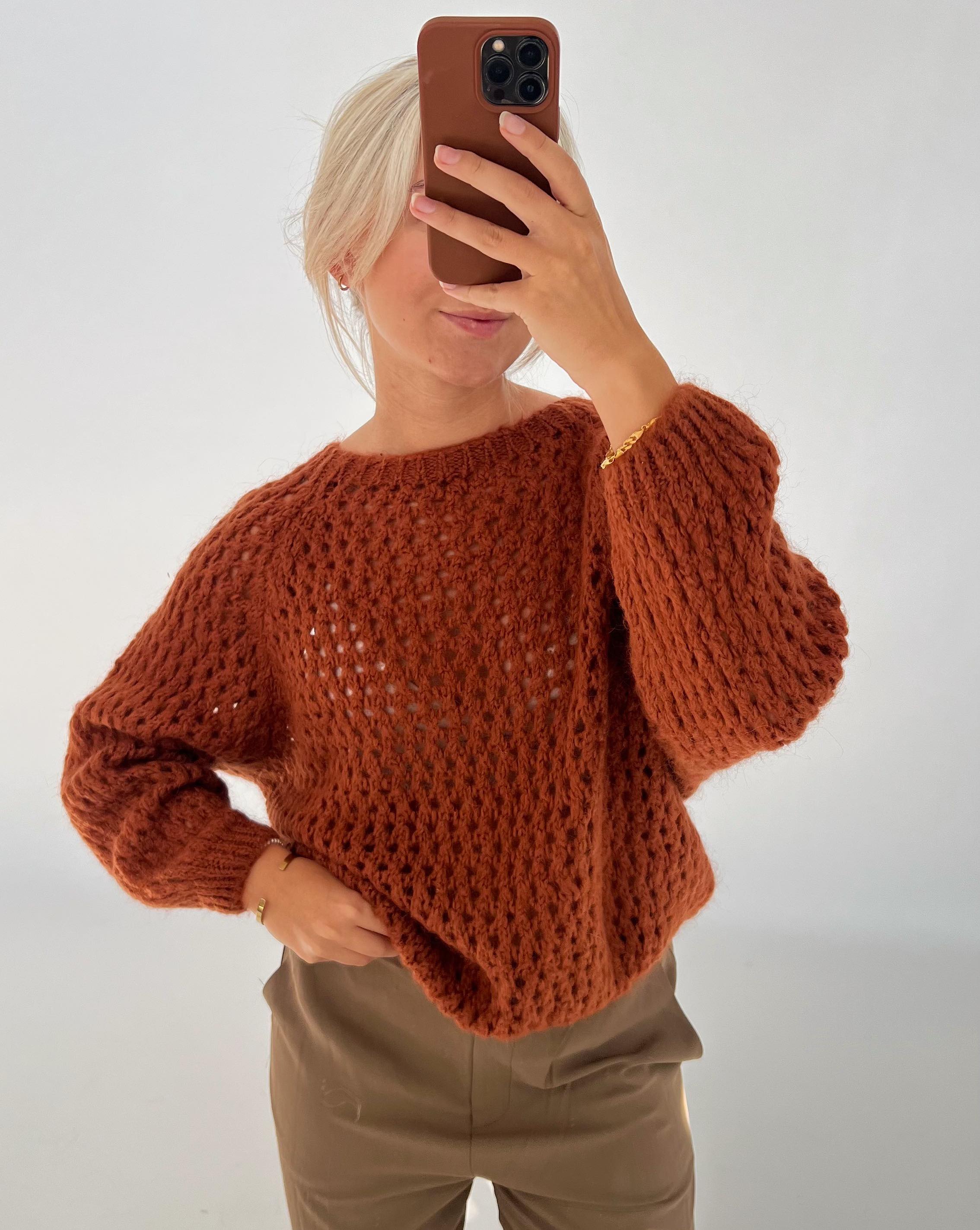 En person iklædt en brun strikket sweater tager en selfie med en mobiltelefon i et lyst rum. De har lyst hår og bærer brune bukser.