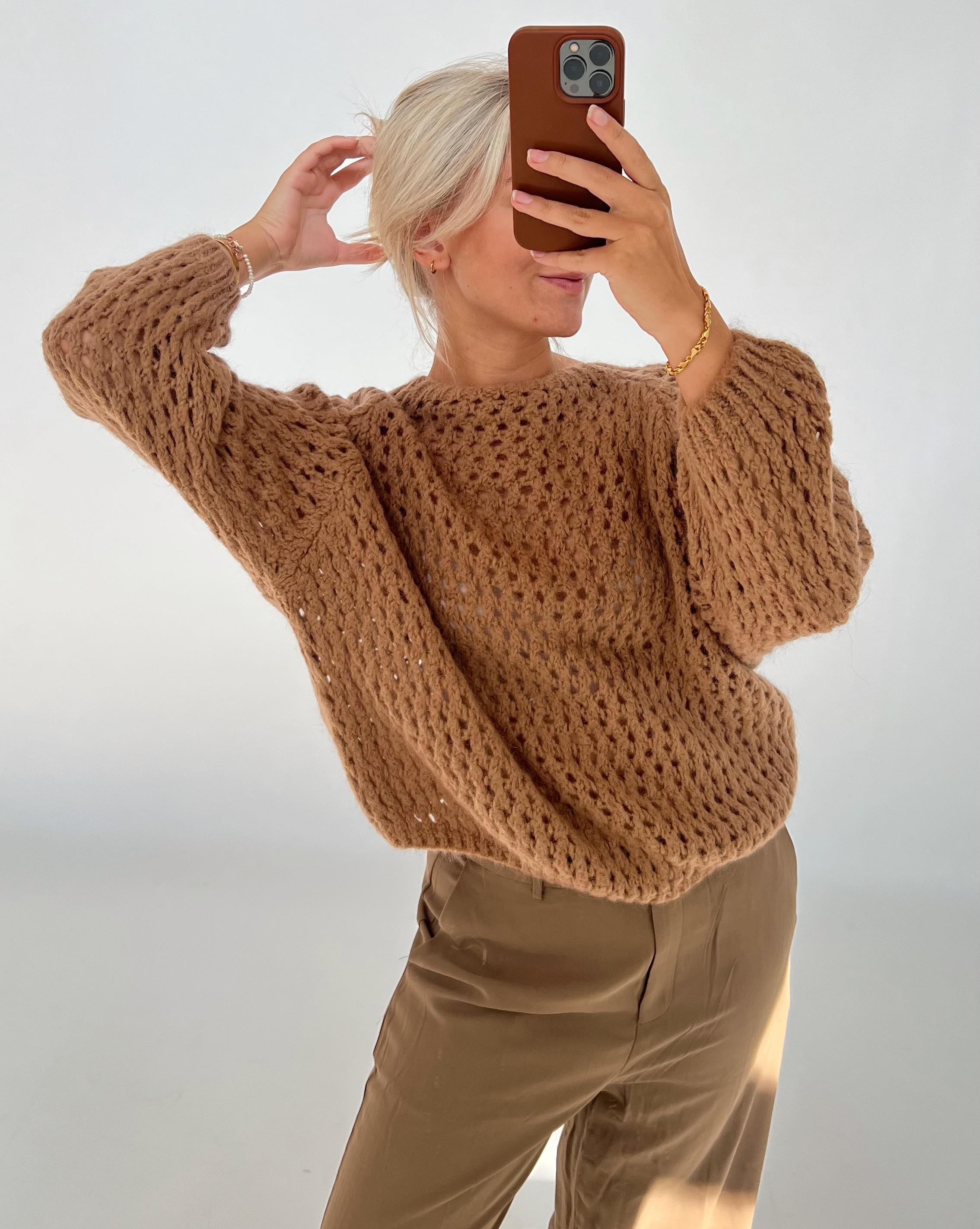 En person tager et spejlfoto iført en brun strikket sweater og beige bukser. Personen holder en telefon op foran ansigtet i et lyst rum.