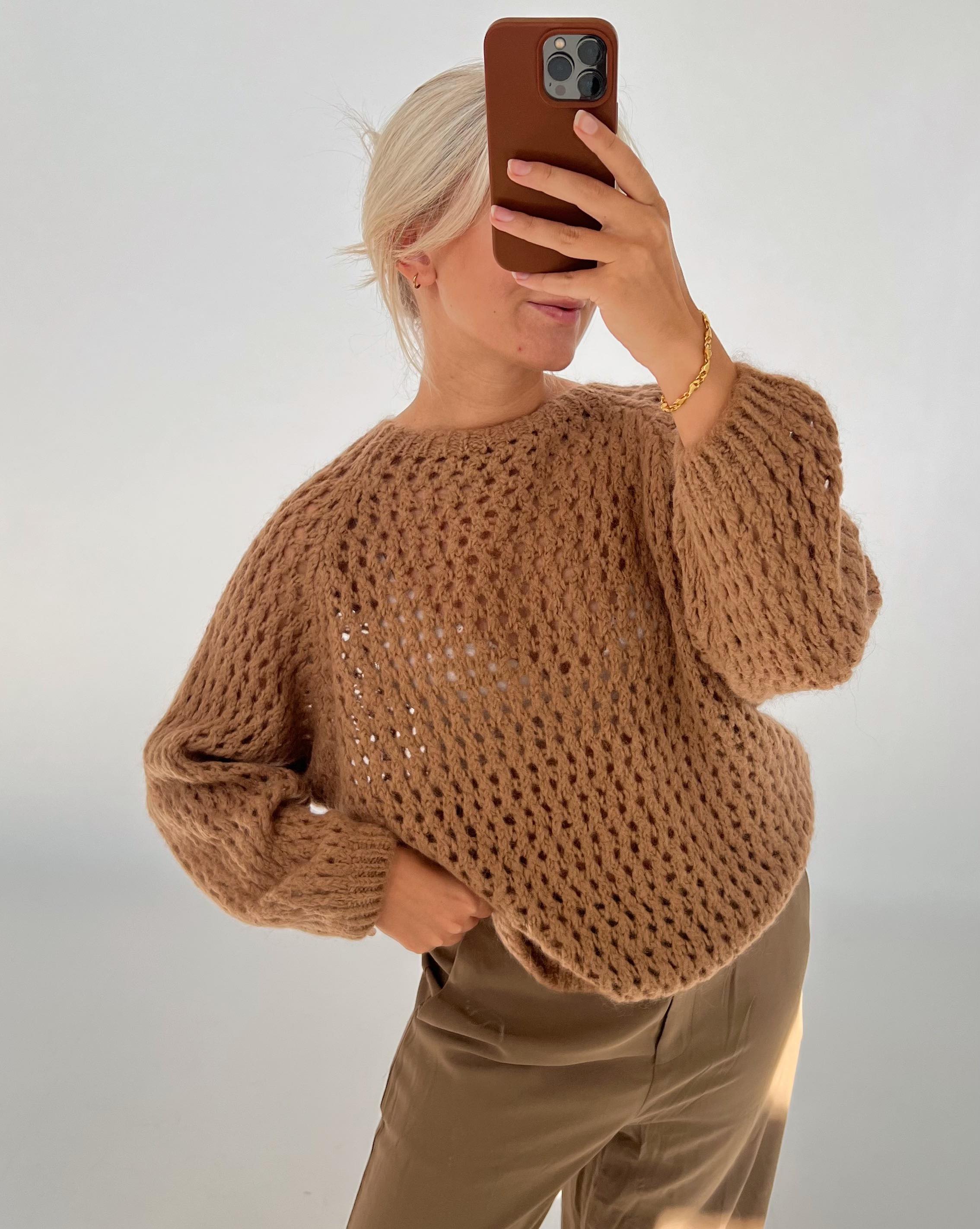 Person i lys brunt, strikket trøje tager et spejlfoto med mobiltelefon. Personen har lyst hår, iført beige bukser, står i et minimalistisk oplyst rum.