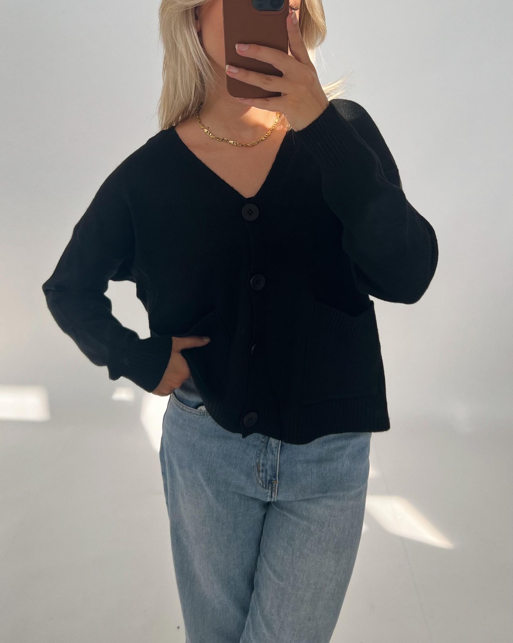 En person iført sort cardigan og lyse jeans tager et spejlfoto. Personen dækker ansigtet med en telefon, og står i et lyst rum.