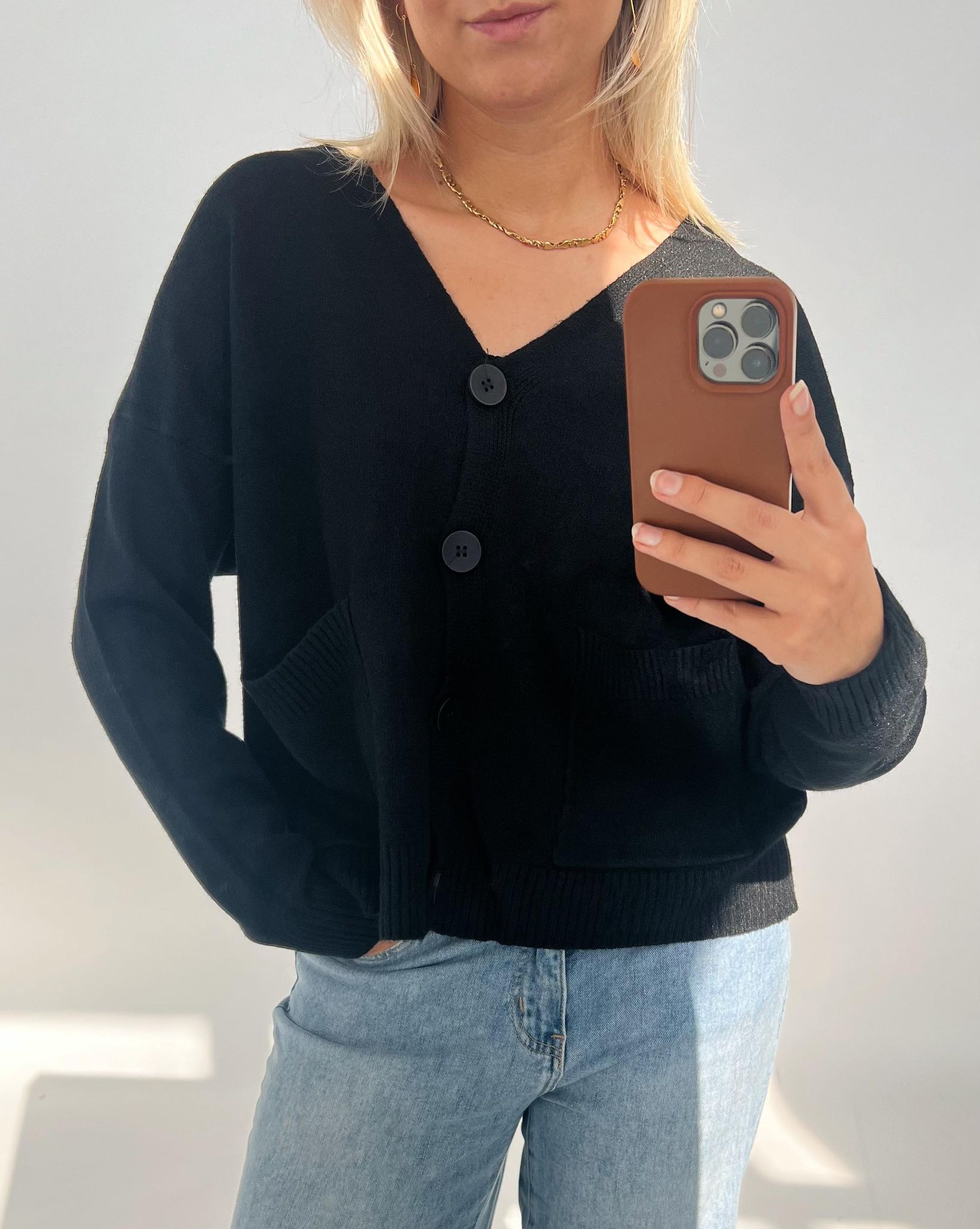 En person iført en sort cardigan med store knapper tager en selfie. De holder en telefon med brunt cover og står i et lyst rum.