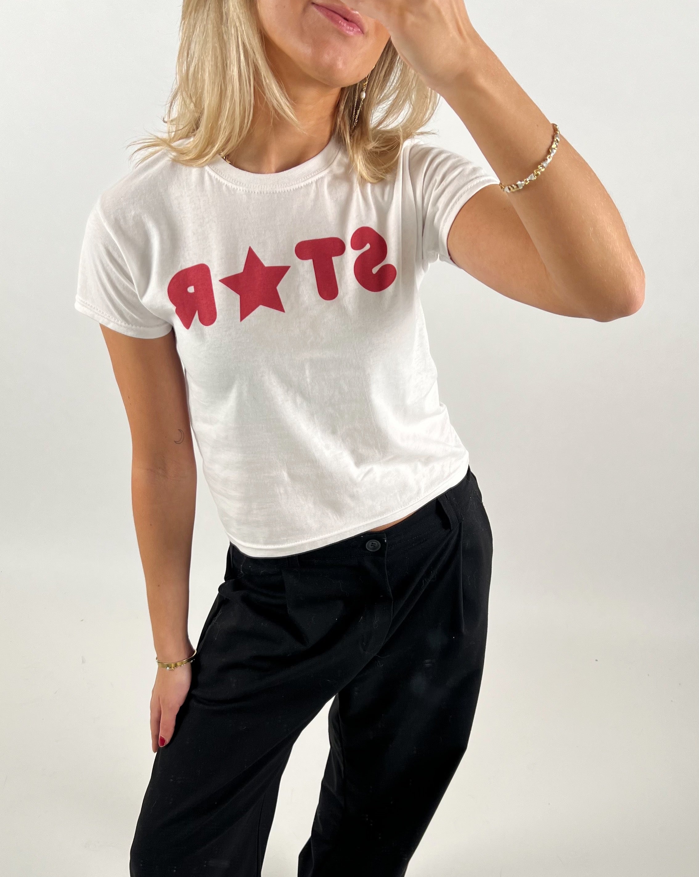 En person iført en hvid T-shirt med rød tekst "STAR," poserer med hånden løftet til ansigtet i en lys indendørs setting. Personen har sort bukser på.