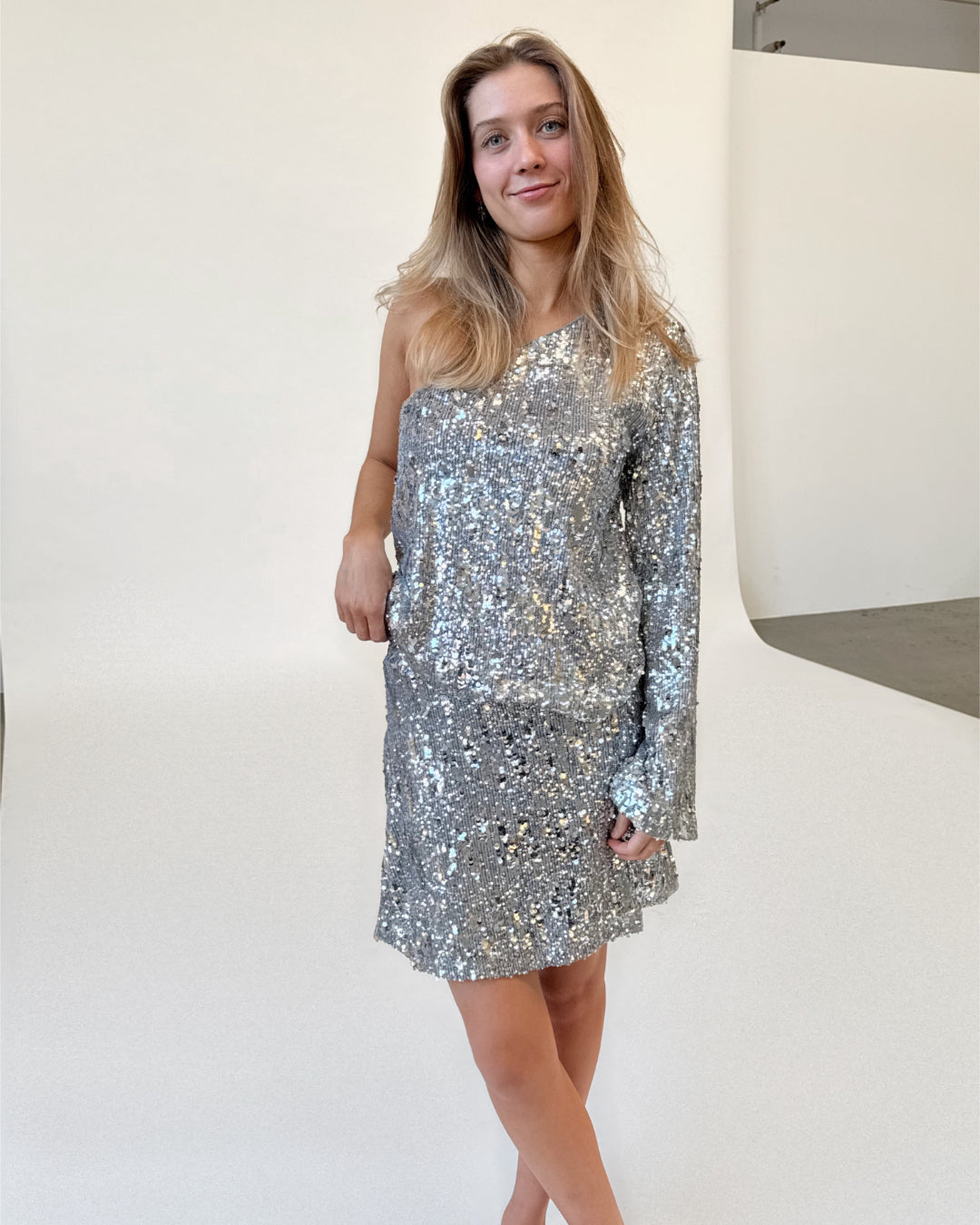 Modelfoto af pige i sølvfarvet TARA nederdel og TARA one-shoulder kjole med pailetter. Alt er fra BySofieSønderby.