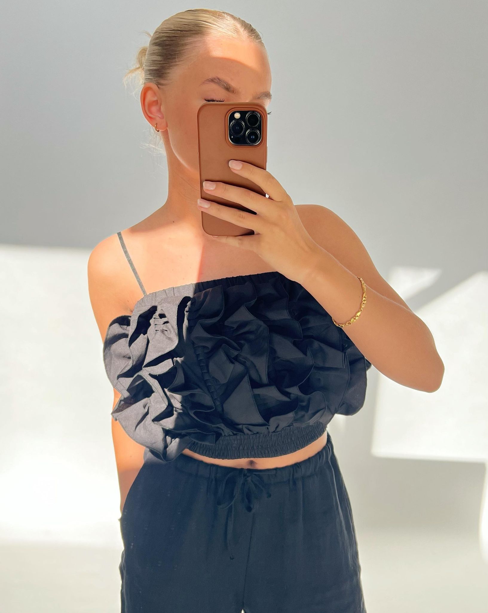Person tager selfie, iført sort top med flæser, holder brun telefon foran ansigtet i lyst rum. Arm bærer gyldent armbånd. Ingen tekst til stede.