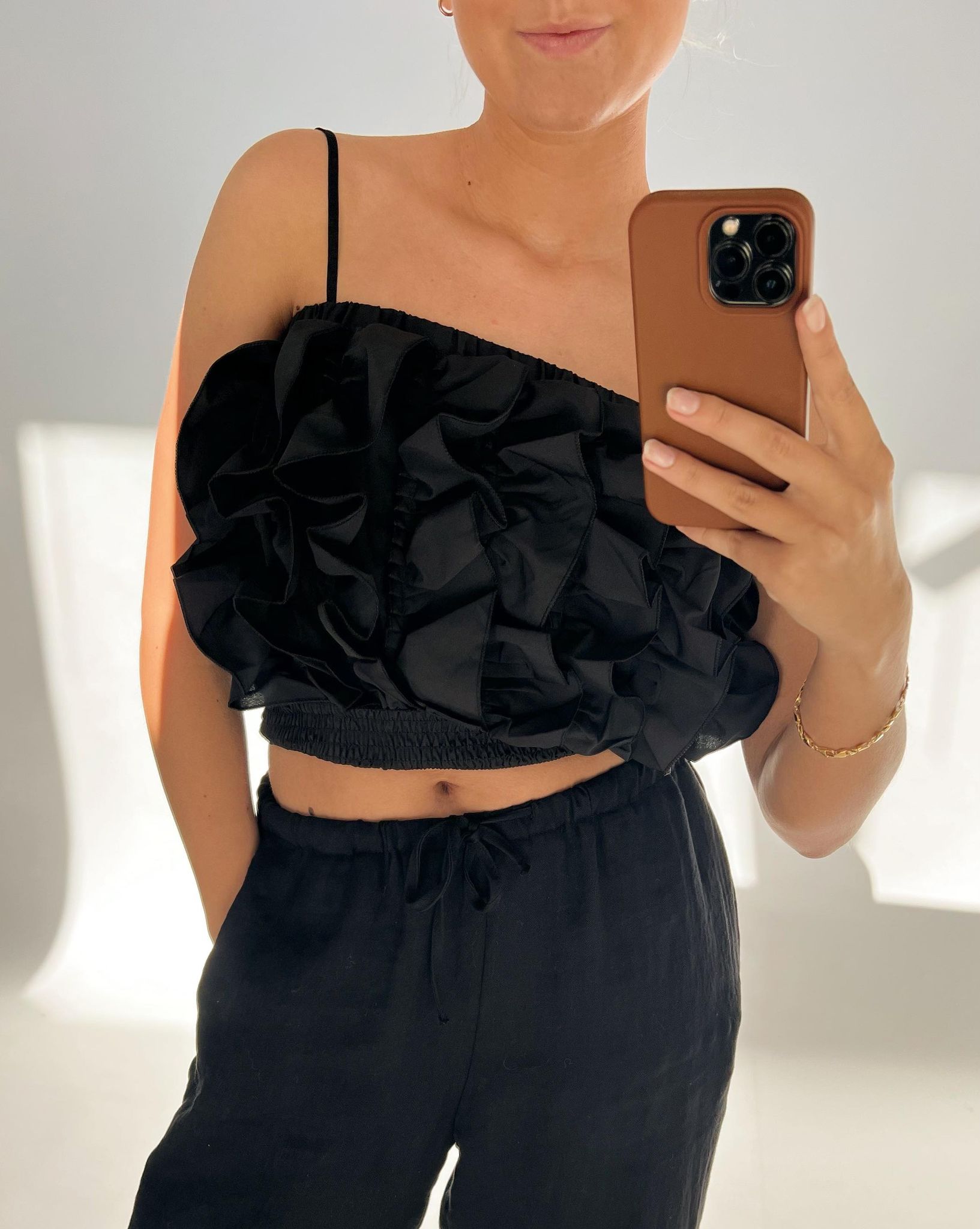 En person iført sort rynket top og bukser tager en selfie med en brun mobiltelefon i et lyst rum. Personen bærer et armbånd og står med ene hånd i lommen.